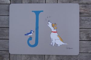 This placemat has a background the colour of unbleached linen on which sits an illustrated blue capital letter J, decorated with a Jack Russell who is singing with his friend the Jay bird. music notes are sketched in above his head.