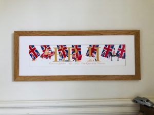 A framed print created to celebrate Queen Elizabeth II's platinum Jubilee, showing her name ERII in large gold lettering with a back drop of large union jack flags infront of which parade mounted and on foot soldiers seen in the parade