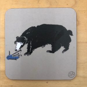 This animal coaster shows a black bear with a white face looking bemused at an object he has found on the ground - some bagpipes. Image on a warm grey melamine background and coaster backed wit cork