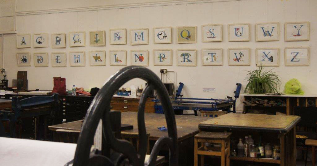 photo of a printmakers' workshop , with a completed A - Z illustrated alphabet displayed on the wall in 26 white frames.