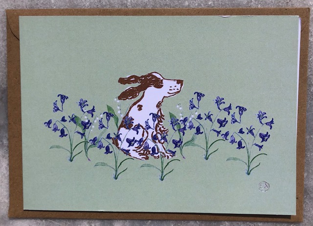 a liver and white springer spaniel sits happily amongst the spring bluebells and lily of the valley, against a light green background.