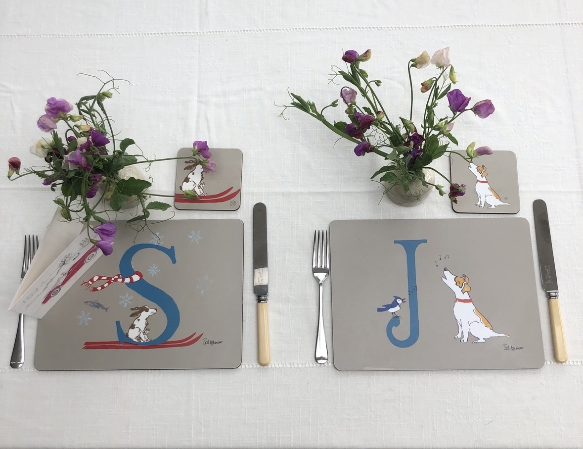 A table setting of 2 illustrated placemats and coasters. One is illustrated with a blue letter S and spaniel on red skis, a scarf and leaping salmon; the other, letter J for a jack russell singing with a jay bird.
