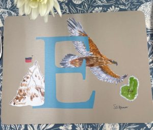 A melamine placemat depicting a large blue letter E illustrated with an eagle in flight who has just planted nautical flag E on Everest and is heading to Eigg, Scotland, home of the golden Eagle